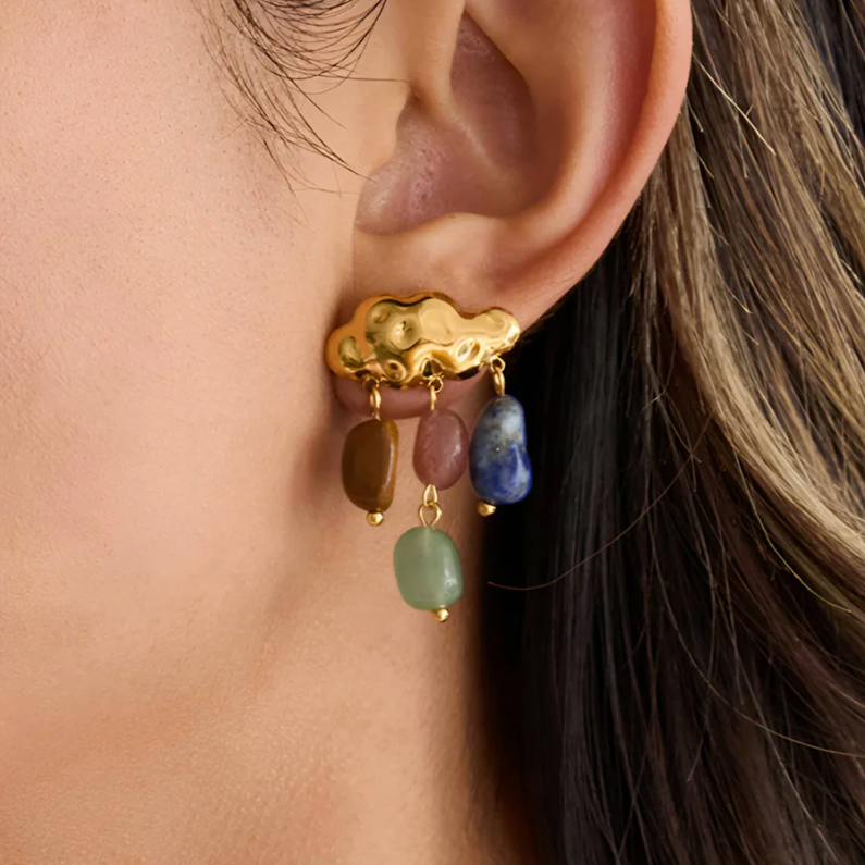 A close-up of a person's ear wearing a gold cloud-shaped stud earring with dangling multicolored gemstone charms.