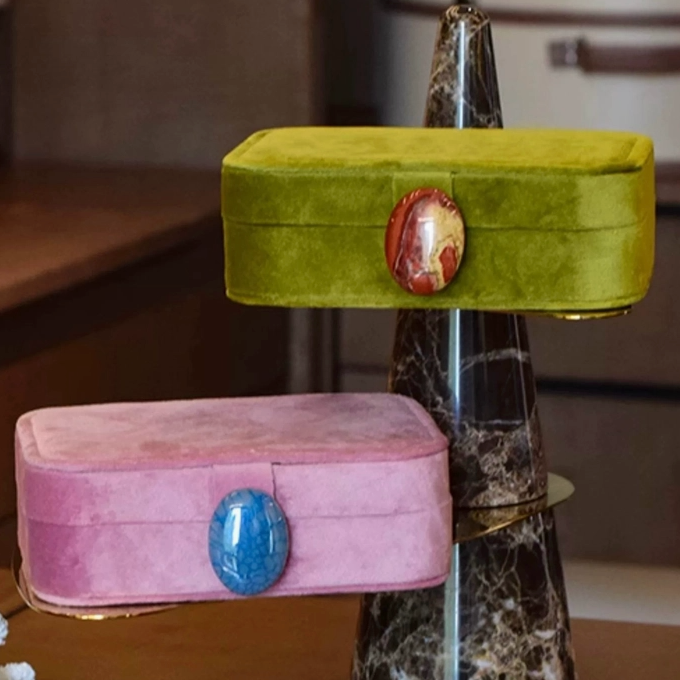 Two velvet jewelry boxes displayed on a marble stand in an indoor setting. The top box is olive green with a round red and gold cabochon, and the bottom box is dusty pink with a blue cabochon.